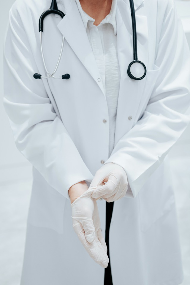 A physician in a white coat with a stethoscope pulling on an exam glove
